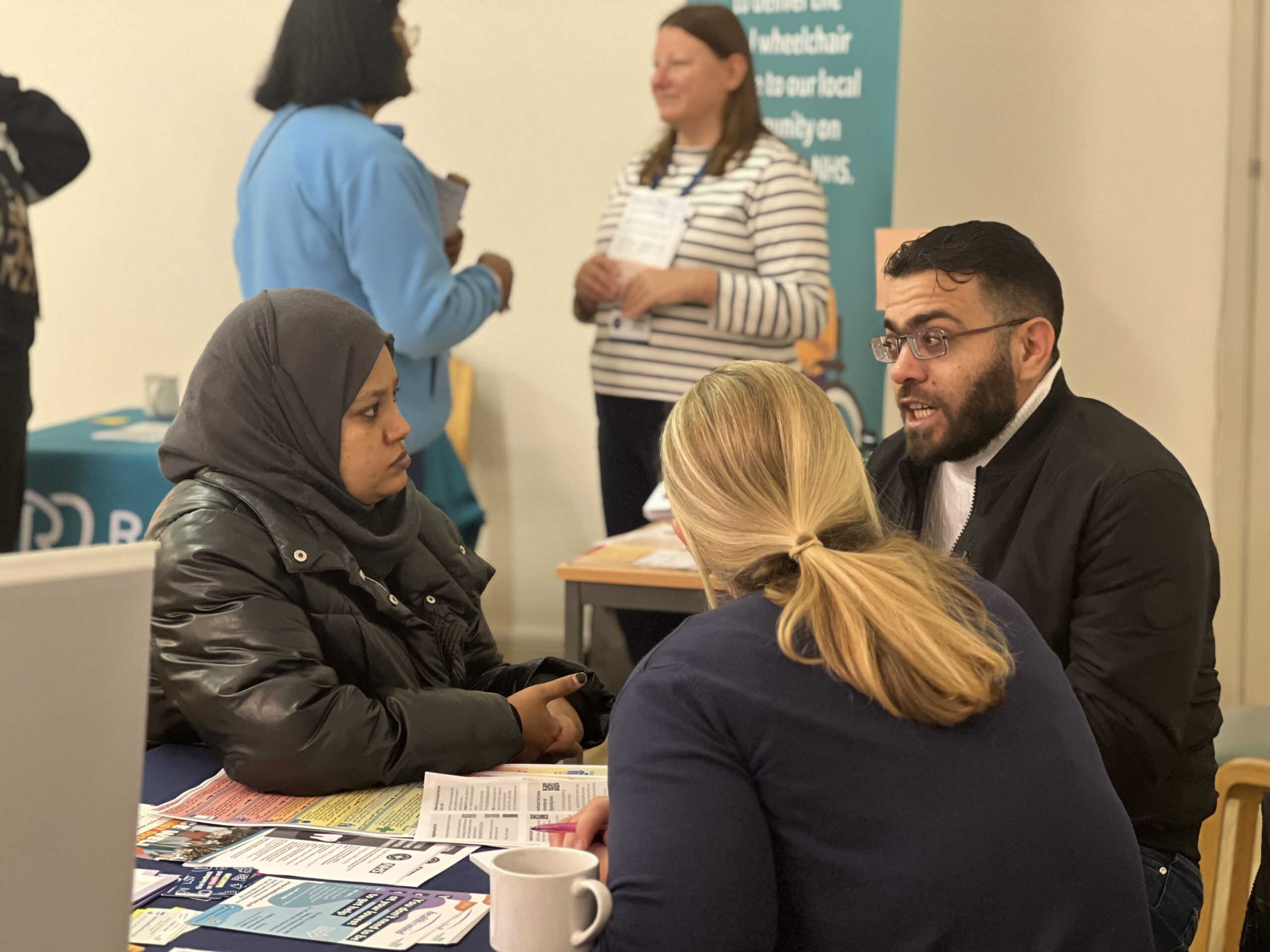 A woman wearing a hijab is on the left, looking at a man who is the talking. The man has a beard and wears glasses. He is talking to a woman with blonde hair. The woman with a hijab is interpreting for him.