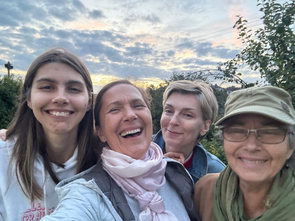 A group selfie of four women smiling and laughing outdoors at sunset in Hastings during their Carers peer-support group meeting. Behind the group is a cloudy sky, and in the background, there's a garden.
