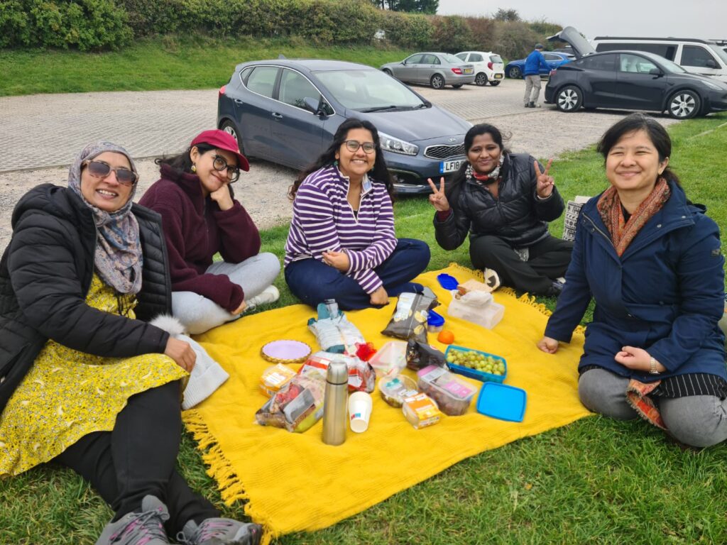 Five women who attended DRI's Women's Walk at Ditchling Beacon are seated on a picnic blanket on a grassy area, smiling at the camera.