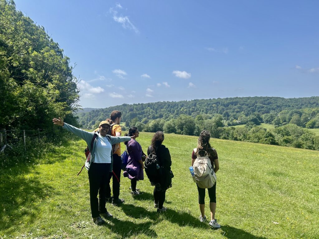 Four individuals enjoy the view of the Sussex Downs from a green hill in Stanmer Park. Mebrak, alongside them. is extending her arms and smiling at the camera.