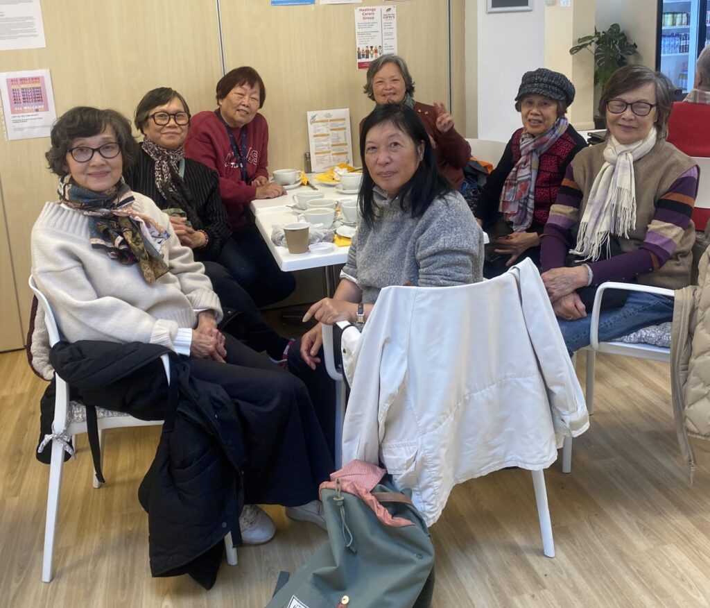 A photo of a group of seven older Hong Kongnese female unpaid carers seated around a white table in a bright, modern community hall in Hastings.