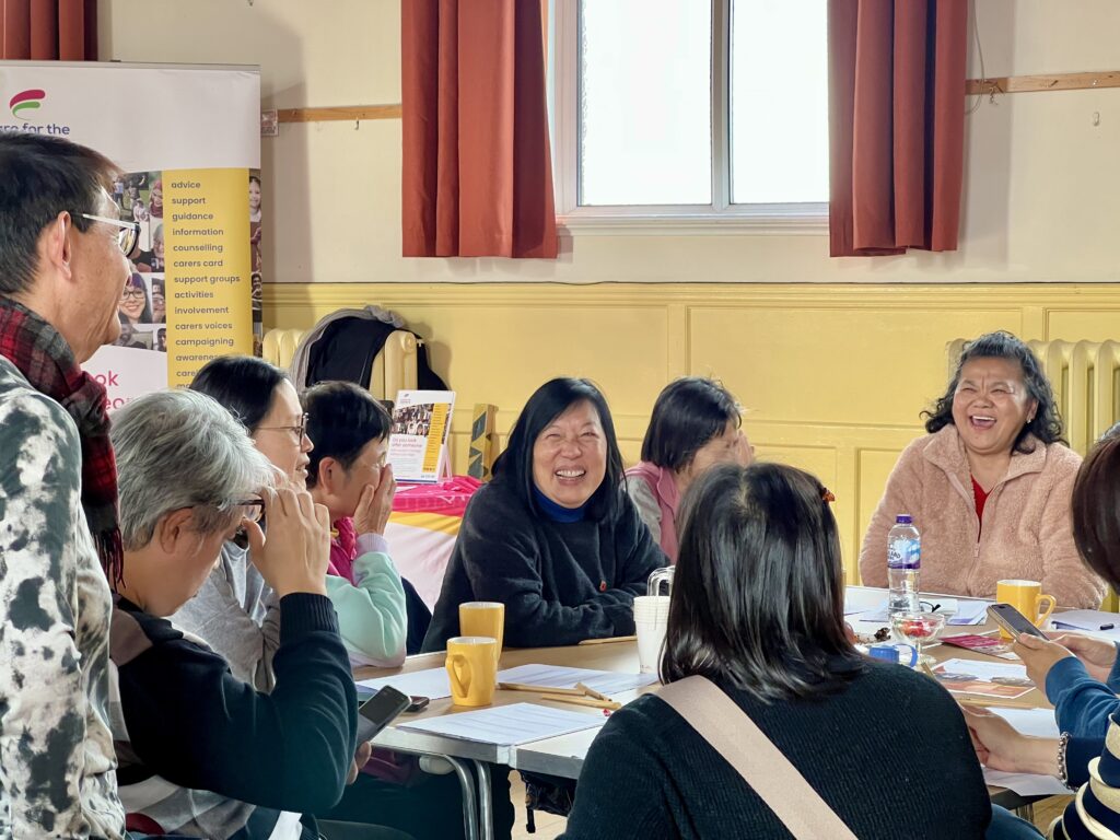 A close-up shot of a group of mostly older Chinese women laughing and engaging in conversation around a table at the Stroke Awareness workshop in Eastbourne. In the center, a woman with dark hair and a black sweater is smiling broadly at the camera. To her right, another woman in a light pink fluffy top is laughing. A man on the far left, partially visible, is also smiling and engaged.