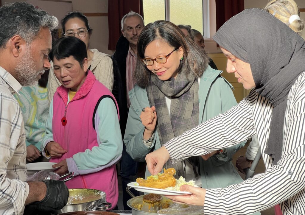 A group of diverse people gathered around a buffet-style serving area at the DRI Stroke Awareness Workshop in Eastbourne, where a man is serving food onto plates. In the foreground, a woman wearing a black hijab and a black-and-white striped shirt holds out a plate toward the man for a portion of a chicken curry dish. Behind them, a woman in a light turquoise jacket and a plaid grey scarf is smiling and looking down at the plate. Several other people of various ages and ethnicities are visible standing in line behind the main action.