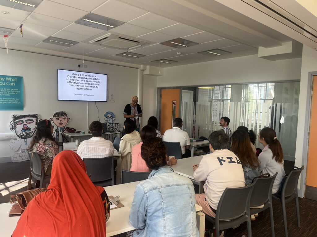 A training session in a bright, modern room. Dr Yaa Asare, a woman with short, grey hair wearing a black and white patterned shirt, is standing near the center, addressing the group.