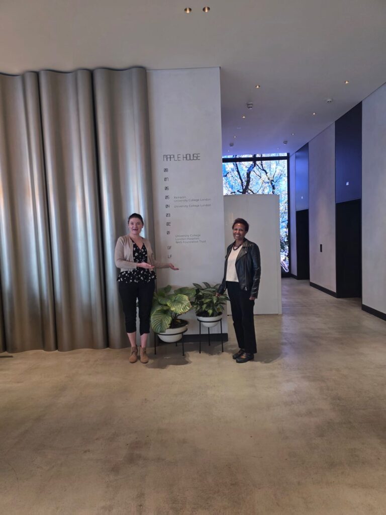 Tacye Turner and Mebrak Ghebreweldi are standing in a lobby. Tacye is gesturing toward a directory sign, which reads "MAPPLE HOUSE" and lists floor tenants, including King's College London and University College London.