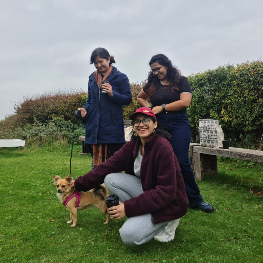 Three participants of one of DRI's Women's Walks are outdoors in a grassy area. One woman is crouching in the foreground, petting a small dog, which is wearing a pink harness. The background features a low hedge of thick green and brown bushes, a rough wooden bench, and a very cloudy, bright sky.
