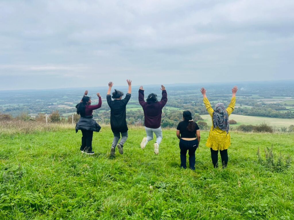Five ethnically diverse women jumping in the air on a grassy hill overlooking a wide, rural valley landscape. Their arms raised above their heads in a celebratory pose. The photo was taken at one of DRI's Women's Walks.