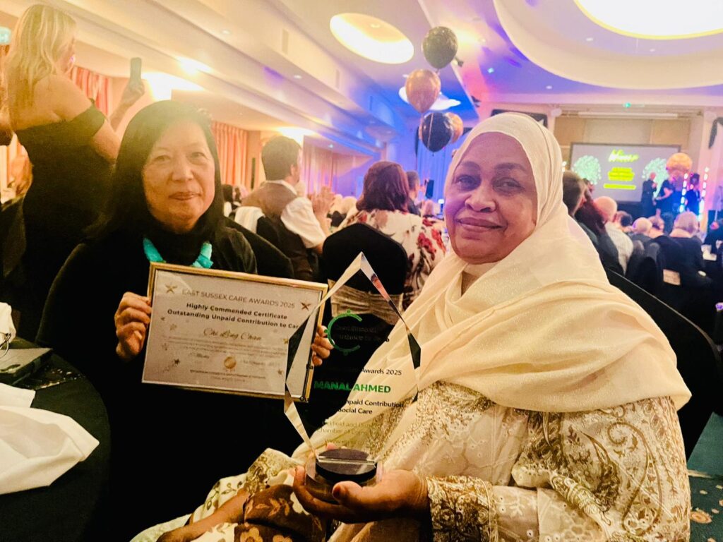 Chi Ling Chan and Manal Ahmed proudly display their awards while at the ESPIC Annual Care Awards ceremony 2025. Manal, wearing a light-colored cream and gold embroidered dress and a matching hijab, is smiling directly at the camera while holding her clear, star-shaped glass award. Chi Ling, with long dark hair and a black top with a turquoise accessory, is holding up her certificate.